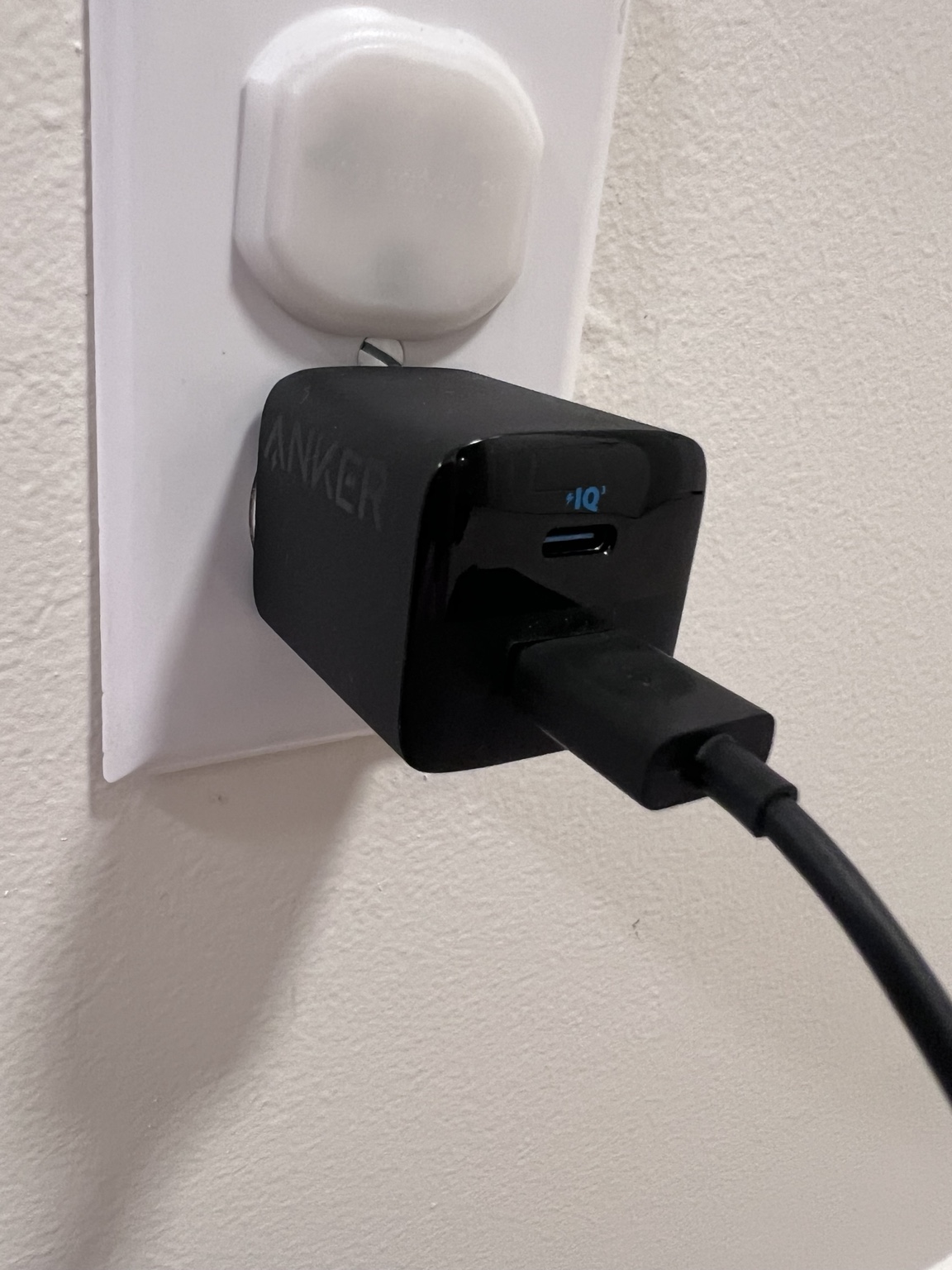 My black Anker wall charger plugged into an outlet, with the Anker logo and USB-C port visible and a USB-A cable connected.
