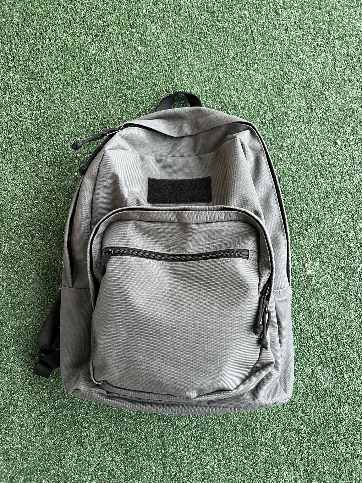 My grey backpack on green ground cover, showing its front zip pockets and black patch panel.