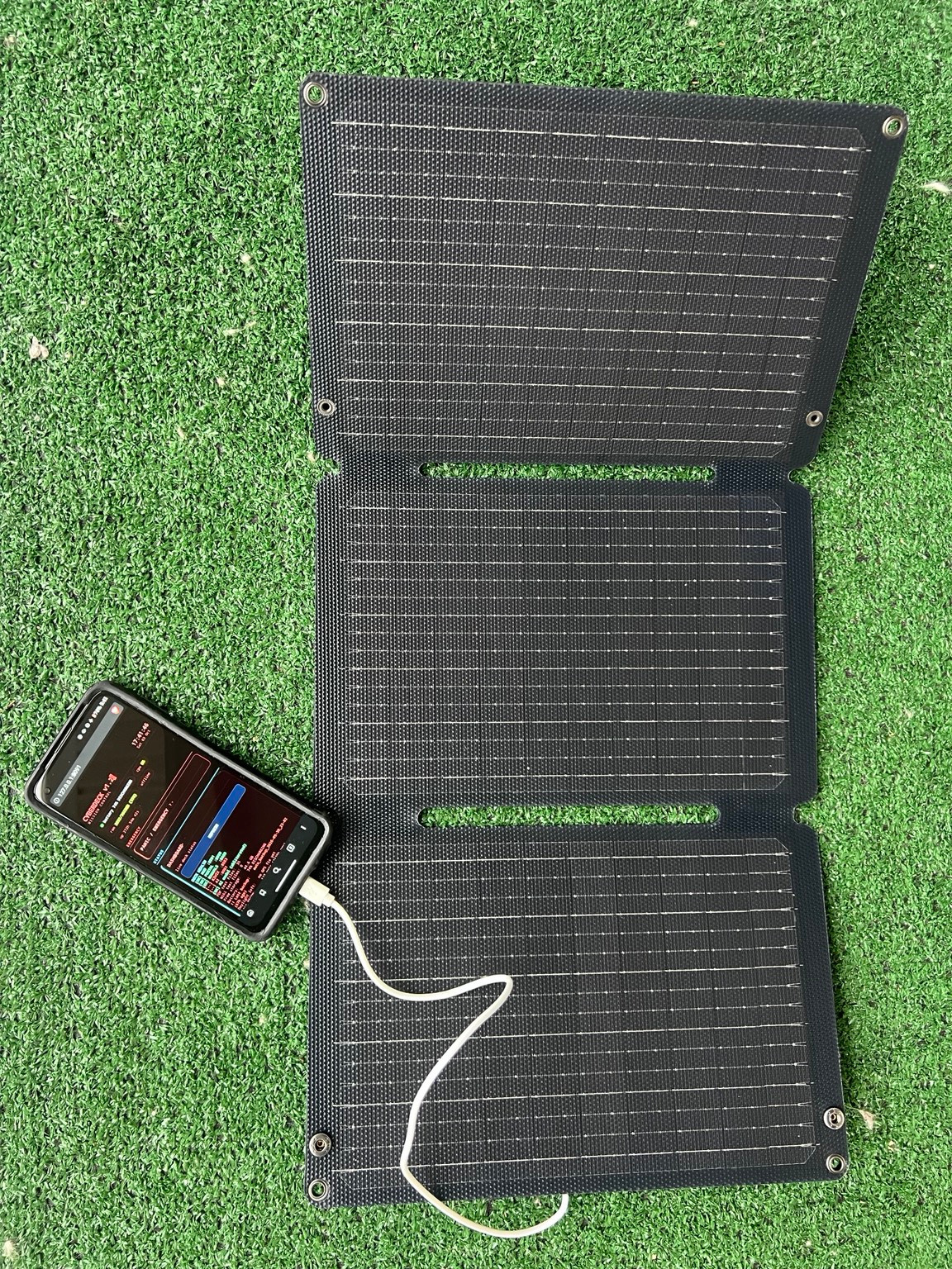 My unfolded three-section BigBlue solar panel on green ground cover, with a cable connected to the cyberdeck phone.