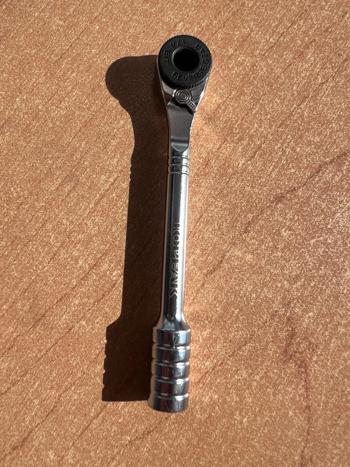 Chrome ratcheting bit driver on a tan surface, showing its round black ratchet head and grooved handle.